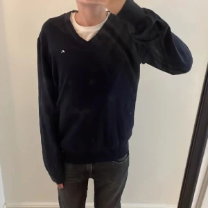 Mörkblå tröja från J.Lindeberg - Snygg mörkblå tröja från J.Lindeberg med v-ringning. Perfekt för en stilren look. Tröjan har långa ärmar och ribbade muddar vid ärmslut och nederkant.