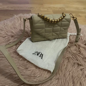 Beige quiltad axelväska från Zara - Snygg beige axelväska från Zara med quiltat mönster och guldfärgade pärldetaljer på handtaget. Väskan har en justerbar axelrem och en praktisk dragkedja för säker förvaring. Perfekt för att ge en touch av elegans till din outfit.