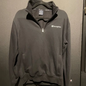 Halv-Zip tröja från Champion - Säljer en svart tröja från Champion med halv dragkedja och broderad logga på bröstet. Tröjan har långa ärmar och en hög krage, perfekt för kyliga dagar. Den är tillverkad i ett mjukt material för extra komfort.