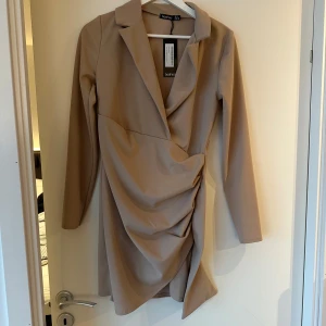 Beige omlottklänning från Boohoo - Snygg beige omlottklänning från Boohoo med långa ärmar och draperad design. Perfekt för en stilren look med en elegant touch. Klänningen har en klassisk krage och är i en smickrande kort längd.