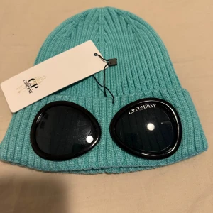 Turkos ribbad mössa från C.P. Company - Snygg turkos mössa från C.P. Company med ribbad design och inbyggda glasögon. Perfekt för att sticka ut i mängden och hålla sig varm. Glasögonen är svarta och pryds av C.P. Company-loggan.