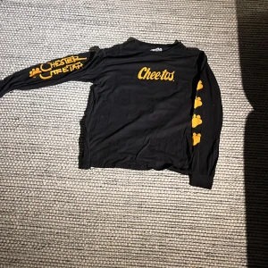 Svart långärmad Cheetos-tröja - Cool svart långärmad tröja med Cheetos-tryck i gult. Framsidan har texten 'Cheetos' och baksidan visar Chester Cheetah som dunkar en basketboll. Tillverkad i 100% bomull, perfekt för avslappnade dagar.