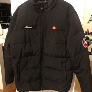 Svart pufferjacka från Ellesse - Snygg svart pufferjacka från Ellesse med dragkedja och broderad logga på bröstet. Perfekt för kyliga dagar med sin quiltade design och långa ärmar. Jackan har en klassisk look och är lätt att matcha med olika outfits.