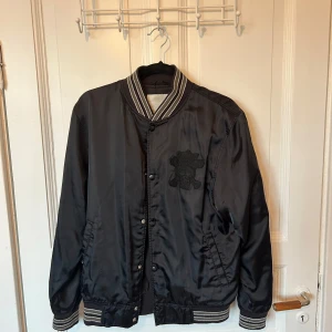 Svart bomberjacka från Stussy - Snygg svart bomberjacka från Stussy med broderad logga på bröstet och stort Stussy-tryck på ryggen. Jackan har knappar framtill och randiga detaljer på krage, ärmslut och nederkant. Det är en vintage jacka vilket också innebär att den är några år gammal, men den har varit väl omhändertagen och är i bra skick.