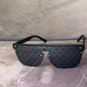 Louis Vuitton Waimea - Stilrena svarta solglasögon från Louis Vuitton med monogrammönster på glasen. Tillverkade i Italien, dessa glasögon har en modern och edgy design med en bred ram och subtila detaljer. Perfekta för att ge en lyxig touch till din stil. OBS kan ej garantera att dom är helt äkta!