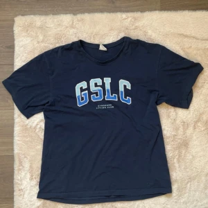 Mörkblå t-shirt från Gymshark - Säljer en mörkblå t-shirt från Gymshark med trycket 'GSLC' i vitt och blått på framsidan. T-shirten är kortärmad och tillverkad i bomull, perfekt för en avslappnad stil.