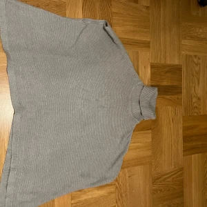 Grå stickad poncho - Säljer en stilren grå stickad poncho med polokrage. Perfekt för kyliga dagar och enkel att matcha med olika outfits. Ponchon har en lös passform och är ett mångsidigt plagg för garderoben.