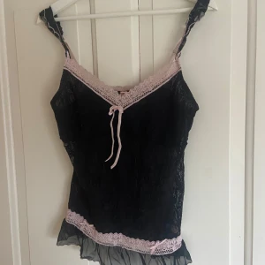 Svart spetslinne från Sophie B. - Snyggt svart spetslinne från Sophie B. med rosa spetsdetaljer och knytband framtill. Perfekt för en elegant look med sina tunna axelband och volangkant nedtill. Passar perfekt som fest topp eller pyjamas. Köpt i new york. Är i large men passar bra på m, eventuellt small. Pris kan diskuteras