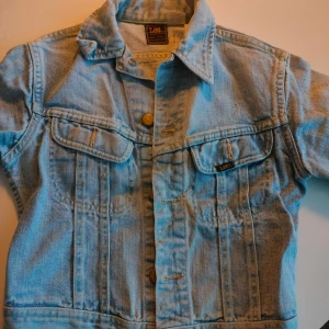 Ljusblå jeansjacka från Lee - Snygg ljusblå jeansjacka från Lee med klassisk krage och knappar framtill. Jackan har två bröstfickor med knappstängning och märkets logga på ena fickan. Perfekt för en avslappnad stil.