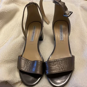 Bronsfärgade sandaler från Sandra Morales - Eleganta bronsfärgade sandaler från Sandra Morales med en stilren design. Skorna har en justerbar rem runt ankeln och en öppen tå. Perfekta för en somrig look.