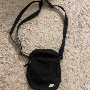 Svart axelväska från Nike - Snygg och praktisk svart axelväska från Nike med justerbar rem och dragkedja. Perfekt för att bära med sig det nödvändigaste på ett stilrent sätt. Väskan har en enkel design med Nike-loggan på framsidan.