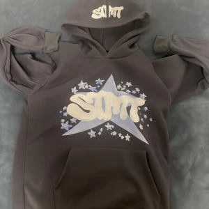 Grå hoodie med stjärnmönster - Snygg grå hoodie med en stor stjärna och texten 'SMT' i vitt på framsidan. Hoodien har en rymlig ficka framtill och en bekväm huva med samma textdetalj. Perfekt för en avslappnad stil.