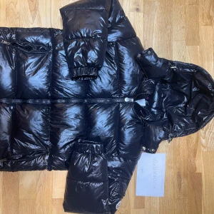 Moncler Maya Jacka - Snygg svart dunjacka från Moncler med glansig finish och praktisk huva. Jackan har dragkedja och långa ärmar, perfekt för kyliga dagar. Den är stilren och håller dig varm hela vintern.