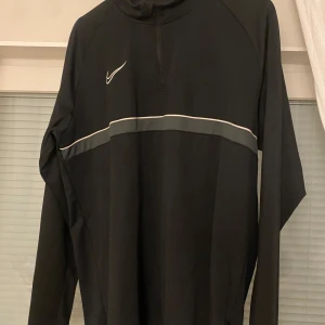 Dryfit Nike - Säljer en svart långärmad tröja från Nike med en vit logga på bröstet och en grå rand över bröstet. Tröjan har en halv dragkedja vid halsen och är perfekt för sportiga aktiviteter.