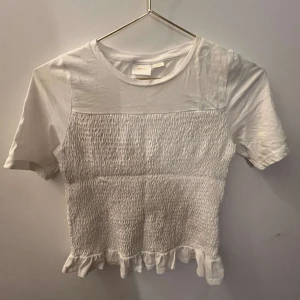 Vit topp från Gina Tricot - Säljer en söt vit topp från Gina Tricot med korta ärmar och smockdetaljer på framsidan. Toppen har en volangkant nedtill som ger en extra touch. Perfekt för en avslappnad stil.