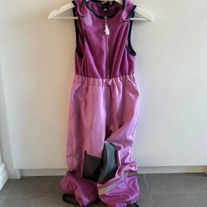 Lila byxdress för barn - Säljer en lila byxdress för barn med dragkedja framtill. Byxdressen har förstärkta knän i mörkare nyans och elastiska benslut. Perfekt för lek och utomhusaktiviteter.