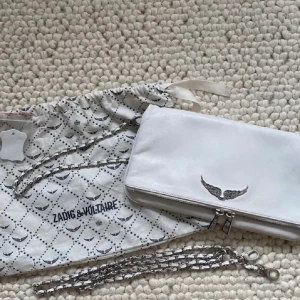 Vit axelväska från Zadig & Voltaire - Snygg vit axelväska från Zadig & Voltaire med silverdetaljer och en kedjerem. Väskan har en del slitage därav säljer jag den billigt, svårt att få med slitaget på bild men skriv för mer bilder 