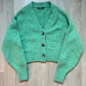 Grön stickad kofta Gina Tricot - Mysig stickad kofta från Gina Tricot. Koftan har en v-ringad design med knappar framtill och ribbade muddar vid ärmslut och nederkant. Aldrig använd!