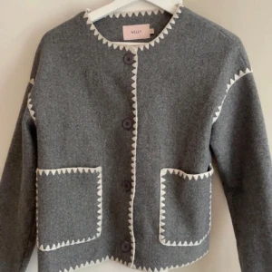 Grå kappa från Nelly - Snygg grå kappa från Nelly som inte går att köpa längre, den har vita zigzag-detaljer längs kanterna. Kavajen har stora fickor framtill och knäpps med knappar. Perfekt för en vår/höst look.