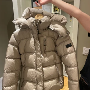 Beige dunjacka från Cedrico - Nypris 6000kr! Säljer en snygg beige dunjacka från Cedrico med hög krage och avtagbar huva. Jackan har en dragkedja framtill och flera praktiska fickor. Perfekt för kalla dagar!