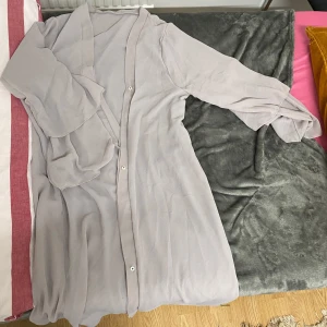 Ljusgrå abaya med knappar - Elegant ljusgrå abaya  med knappar framtill och långa ärmar. Klänningen har en v-ringad design och är perfekt för en stilren look.