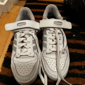 Vita sneakers från Adidas - Säljer ett par vita Adidas sneakers med snörning och kardborreband. Skorna har de klassiska tre ränderna på sidan och perforerade detaljer på tån för extra ventilation. Perfekta för en sportig look! Väldigt bra skick, dom ser typ smutsig ut men de e ljuset! Knappt använt