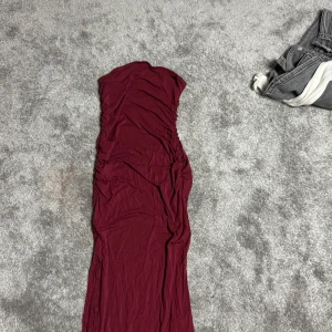 Vinröd långklänning - Elegant vinröd långklänning med en smickrande passform. Perfekt för festliga tillfällen. Klänningen är axelbandslös och har en stilren design.
