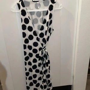 Vit och svart prickig klänning - Snygg ärmlös klänning med stora svarta prickar på vit bakgrund. Klänningen har en v-ringad design och ett knytband i midjan för en smickrande passform. Perfekt för en stilren look.