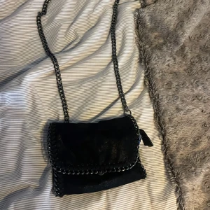 Svart axelväska med kedjedetaljer - Snygg svart axelväska med kedjedetaljer och dragkedja. Väskan har en röd insida och en praktisk storlek för vardagsbruk. Perfekt för att ge en edgy touch till din outfit.