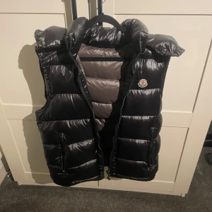 Svart dunväst från Moncler - Säljer en snygg svart dunväst från Moncler. Västen har en glansig finish och är perfekt för kyligare dagar. Perfekt för höst, tags finns och nfc funkar. Storlekn är Y-S vilket passar storlek EU S alldeles utmärkt.