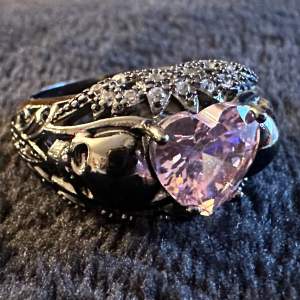 Den här ringen har allt - glittrande cubic zirconerer och en rosa hjärtformad kristall omgiven av dödskallar. 925 sterling silver. Ca 1,8 cm i innerdiameter. Helt makalöst unik, absolut nyskick. Dark romance Säljs då jag inte använder ringar längre. 