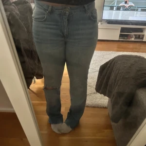 Blå jeans med slitningar - Säljer ett par snygga blå jeans med slitningar vid knäna. De är tighta och har en klassisk femficksdesign. Perfekta för en casual look!