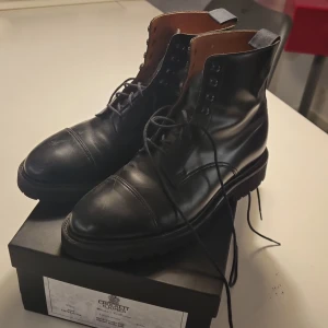 Svarta boots från Crockett & Jones - Säljer ett par stilrena svarta boots från Crockett & Jones. De är i skinn och har en klassisk snörning framtill. Skorna har en robust sula från Vibram, perfekt för höst och vinter. Insidan är fodrad i en ljusare nyans för extra komfort. Perfekta för både vardag och mer formella tillfällen.