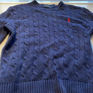 Mörkblå stickad tröja från Polo Ralph Lauren - Säljer en snygg mörkblå stickad tröja från Polo Ralph Lauren. Den har ett klassiskt flätmönster och en liten röd broderad logga på bröstet. Perfekt för höst och vinter, med långa ärmar och ribbade kanter. Supermysig och stilren! Väldigt bra skick.😁