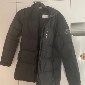 Svart dunjacka från Sail Racing - Säljer en svart dunjacka från Sail Racing i storlek XL. Jackan är perfekt för kalla vinterdagar med sin varma och vadderade design. Den har en dragkedja framtill och en praktisk huva. Jackan är i bra skick och redo för nya äventyr!