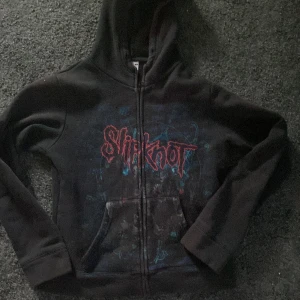 Svart Slipknot hoodie från Fruit of the Loom - Säljer en svart hoodie med Slipknot-tryck från Fruit of the Loom. Den har en dragkedja framtill och en stor ficka. Baksidan pryds av ett stort Slipknot-motiv i rött och blått. Perfekt för fans av bandet! 🎸