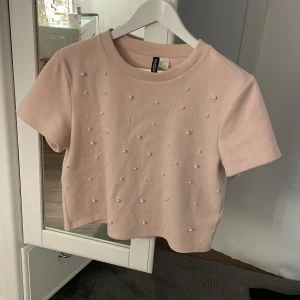 Rosa kortärmad topp med pärlor från H&M Divided - Säljer en söt rosa kortärmad topp från H&M Divided. Toppen är dekorerad med små pärlor på framsidan och har en rund halsringning. Perfekt för en stilren och chic look. 💖