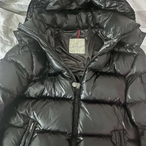 Svart dunjacka från Moncler - Säljer en snygg svart dunjacka från Moncler med glansig finish. Jackan har en dragkedja framtill och praktiska fickor med knappar. Perfekt för kyliga dagar med sin varma vaddering och stilrena design.