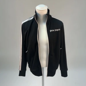 Svart track jacket från Palm Angels - Snygg svart track jacket från Palm Angels med vita ränder längs ärmarna och dragkedja framtill. Jackan har en hög krage och ribbade muddar vid ärmslut och nederkant. Perfekt för en sportig look!