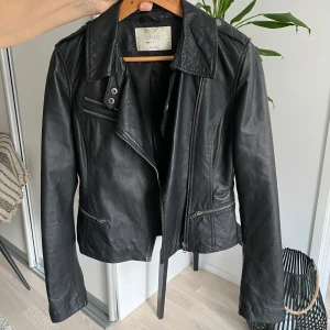 Svart skinnjacka från Only - Snygg svart skinnjacka från Only med dragkedja och knappar. Jackan har en klassisk bikerstil med långa ärmar och flera fickor med dragkedjor. Perfekt för en cool look!