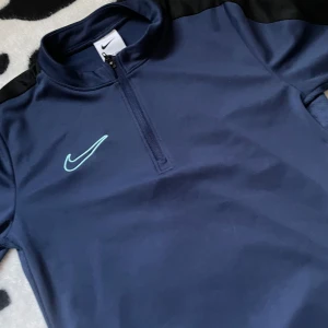 Mörkblå träningströja från Nike - Säljer en mörkblå träningströja storlek ungefär 110-cm från Nike med dragkedja och lång ärm. Perfekt för träning och sportiga aktiviteter. Tröjan har en stilren design med Nike-logga på bröstet. 