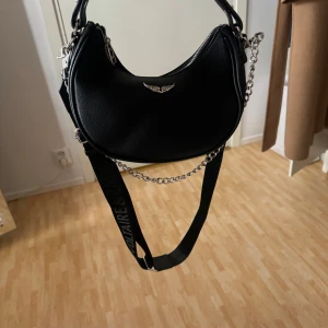 Svart axelväska från Zadig & Voltaire - Snygg svart axelväska från Zadig & Voltaire med coola kedjedetaljer och justerbar axelrem. Väskan har en dragkedja för säker förvaring och är perfekt för att ge din outfit en edgy touch. Perfekt för dig som älskar stil och funktionalitet. Köpt utomlands har ej kvitto, dustbag medföljer 