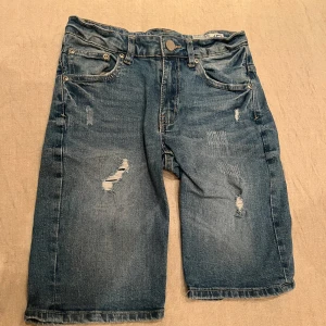 Blå jeansshorts från Shorty Star - Säljer ett par coola blå jeansshorts från Shorty Star. De har en avslappnad passform med slitna detaljer och klassisk femficksdesign. Perfekta för en avslappnad stil.