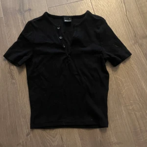 Svart kortärmad topp från Gina Tricot - Säljer en stilren svart kortärmad topp från Gina Tricot. Toppen har knappar framtill och är perfekt för en enkel och snygg look. Passar bra till både jeans och kjol. Perfekt för vardagsbruk!
