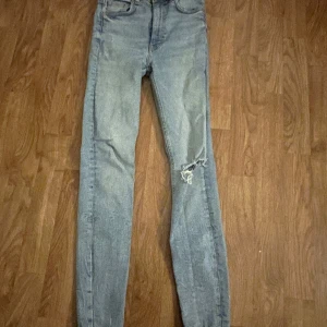 Ljusblå jeans med slitningar - Säljer ett par ljusblå jeans med slitningar på ena benet. De har en skinny passform och är högmidjade. Perfekta för en avslappnad stil.