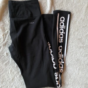 Svarta leggings från Adidas - Snygga svarta leggings från Adidas med Climalite-teknologi. De har en hög midja och ett vitt Adidas-tryck längs benet. Perfekta för träning eller en avslappnad dag.