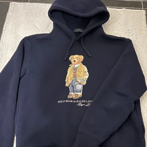 Mörkblå hoodie från Ralph Lauren - Säljer en snygg mörkblå hoodie från Ralph Lauren med en ikonisk Polo Bear-design på framsidan. Hoodien har en klassisk passform med dragsko i huvan och ribbade muddar vid ärmslut och nederkant. Perfekt för en avslappnad stil!
