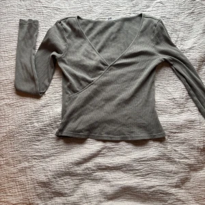 Grön ribbad omlottopp - Säljer en stilren grön ribbad omlottopp med lång ärm. Perfekt för en avslappnad men ändå chic look. Toppen har en v-ringad design som ger en elegant touch. Passar utmärkt till både jeans och kjol.