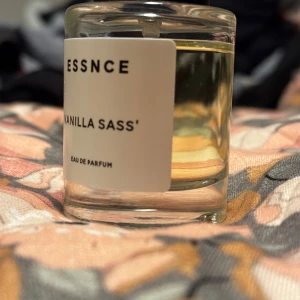 Vanilla Sass' Eau de Parfum - Säljer en flaska Vanilla Sass' Eau de Parfum från Essnce. Parfymen kommer i en stilren, genomskinlig glasflaska med en enkel vit etikett. Doften har en söt och kryddig vaniljton som ger en varm och inbjudande känsla.