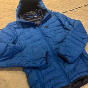 Blå pufferjacka från J.Lindeberg - Snygg blå pufferjacka från J.Lindeberg med dragkedja och huva. Perfekt för kyliga dagar med sin quiltade design som håller värmen. Jackan har en stilren look med en diskret logga på bröstet.